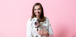 Happy young woman in white jacket cuddling a white and spotted terrier mix dog against a pink backdrop.
