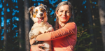 Young woman holding her shiny, happy Australian Shepherd dog outside in the woods on a sunny day